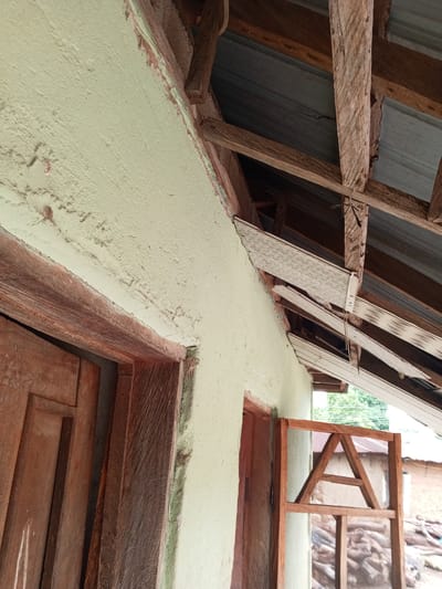 The image shows an unfinished building with exposed wooden roof beams and corrugated metal roofing. The exterior wall is painted a light green, with some areas of rough texture. To the left, a wooden doorframe is visible. To the right, a partially constructed wooden screen with an "A" shape in the top frame is leaning against the wall. In the background, a pile of firewood and another simple structure can be seen, suggesting a rural or semi-rural location. The lighting suggests it is daytime. The scene appears to be in Gindin Dutse, Nigeria, and the building seems to be in a state of construction or repair. There is no visible text in the image.
