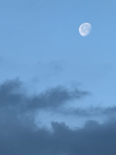 The image shows a blue sky with a waxing gibbous moon visible in the upper right quadrant. The lower half of the image is filled with dark, textured clouds. The scene is devoid of any people, structures, or discernible signs of human activity. The context provided indicates the location is Ol'khovskoye, Russia.