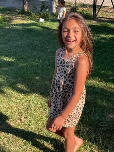 A young girl with long brown hair and a leopard-print dress is smiling at the camera, her legs crossed. She is outdoors on a grassy lawn, with dappled sunlight filtering through trees, suggesting it is daytime. In the background, another child is visible, bending over, and a fence with a metal frame. A small, red and white bucket is on the ground near a bush on the left side of the frame. The scene appears to be a casual, outdoor setting, possibly a backyard or park. The girl's expression is happy and joyful. The location is identified as Oreshak, Bulgaria.