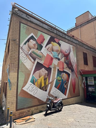 In Naples, Italy, a large, colorful mural covers the side of a brick building. The mural depicts four vintage-style photographs framed by a bird with red and pink feathers. The photographs feature stylized depictions of people in different poses and attire. The top left photo is dated "1930," the top right is dated "1950," the bottom left is dated "1970," and the bottom right is dated "2010." The bird has a camera strapped to its chest. The building's facade is constructed of light brown bricks, with some areas appearing worn. A red railing runs along the top of the building. In front of the mural, on a stone-paved street, rests a silver scooter. Two black bollards with yellow tops and a chain are also present on the street. A small pile of wooden planks lies near the bollards. In the background, parts of other buildings are visible, along with a green storefront with signage and more scooters parked in front. The sky is clear and blue.