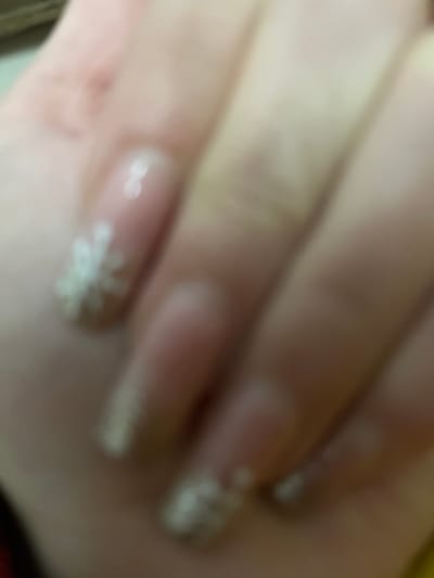 The image depicts a hand with long, manicured nails. The nails are a light pink or nude color, with white snowflake designs at the tips. The overall focus is soft, creating a slightly blurred effect. The hand is positioned at an angle, showing the length and shape of the fingers. The image appears to showcase the nail art.