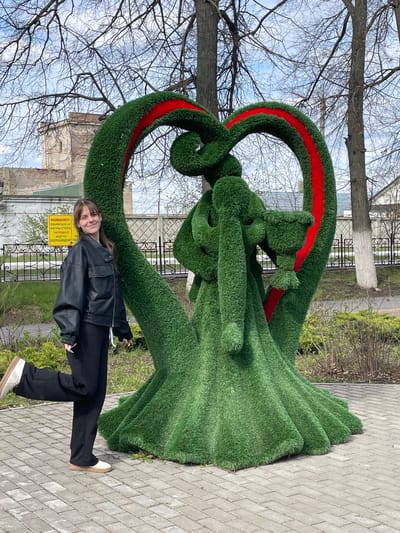 A young woman stands next to a large, green, heart-shaped topiary sculpture with a red stripe on its upper loop. The sculpture depicts a man and a woman in an embrace. The woman wears a black leather jacket, black pants, and white shoes. A yellow warning sign is visible behind the sculpture, attached to a black metal fence. In the background, bare trees reach toward a pale blue sky. The ground is paved with interlocking gray bricks. A building is partially visible in the background, behind the fence. This scene is taking place in the city of Urussu, Russia.