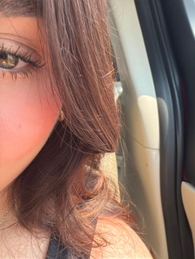 A close-up photograph captures the right side of an individual's face and upper torso, positioned within the interior of a vehicle. The subject's right eye, featuring a hazel or light brown iris and visible eyelashes, is prominently displayed. The cheek exhibits a rosy hue, and the person's long, brown hair with reddish undertones is visible, styled over the right shoulder and partially framing the face. A small, circular, metallic-colored earring is present in the right earlobe, and a thin, metallic chain is partially visible on the upper chest above a dark-colored garment. The background on the right reveals a light-colored vehicle interior door panel with a darker trim element and a faint red accent line. Direct sunlight illuminates the scene from the right, creating highlights and shadows. The image contains no visual identifiers for a specific location within Veracruz, Mexico.