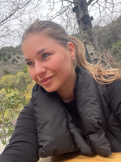 A woman with blonde hair and light eyes is pictured close-up. She wears a black puffer jacket and a black shirt. She has pierced ears. The background includes bare trees and vegetation, suggesting an outdoor location, potentially in a mountainous area. The presence of greenery suggests the season is either late winter or early spring. There is no confirmed information in the image that places this location in Krapina, Montenegro.