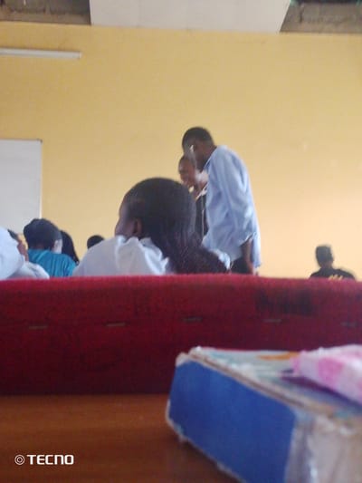 In a classroom setting in Gumel, Nigeria, a group of young people are visible. The walls of the room are painted a light yellow, and a white whiteboard is mounted on the left side. In the foreground, a portion of a red upholstered bench or seating area is prominent. A blue and white box is situated on a wooden surface in the lower right corner of the image.
Several individuals are in the background. A young man wearing a light blue long-sleeved shirt is standing and facing away from the camera, appearing to be engaged in conversation with someone out of frame. To his left, another person, possibly a young woman, is partially visible. In the middle ground, a person with dark hair is seated and facing away. Further to the left, another individual in a blue shirt is also seated. In the far background, a figure is visible wearing a dark shirt, with their head tilted down. The overall lighting suggests an indoor environment, possibly during the day. The image has a watermark in the lower left corner that reads "© TECNO".