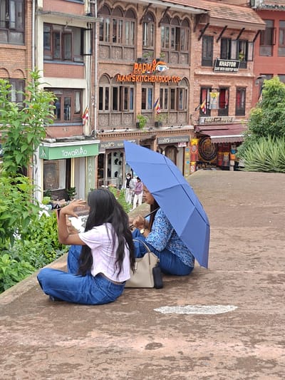 The image captures two women sitting on a weathered, reddish-brown paved surface. The woman on the left is partially visible from the back, with long dark hair, wearing a pink and white tie-dye t-shirt and blue jeans. She is holding a pink smartphone in her left hand and appears to be interacting with it. The woman on the right is facing to the left, holding a large open blue umbrella over both of them. She is wearing a blue patterned long-sleeved shirt and blue jeans. A light-colored handbag rests on the ground next to her. In the background, there is a multi-story brick building with traditional Nepali architectural elements, including intricately carved wooden window frames. Several signs are visible on the building, including "PADMA HOTEL NANI KITCHEN ROOFTOP" and "KHASYOR CAFE WITH ROOFTOP VIEW," indicating this is a commercial area. To the left of the building, a green awning with the text "TAWARiAo" is visible, suggesting a shop. The environment appears to be outdoors, possibly a rooftop or elevated viewpoint in the city of Gokarneshwar, Nepal, given the architectural style and signage. Lush green foliage is present on the left side of the image, framing the scene.