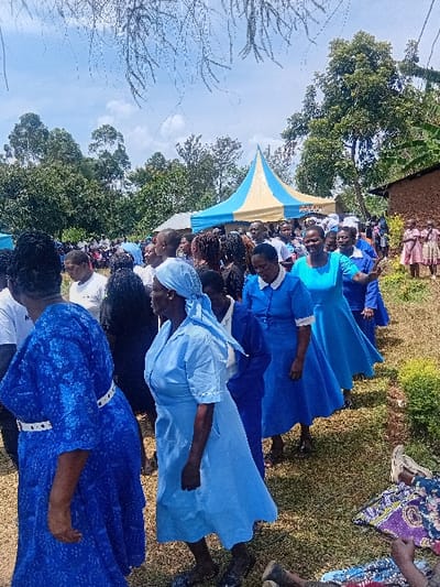 A large group of individuals is gathered outdoors in Butali Chegulo ward, Kenya, under a bright sky with scattered clouds. The foreground shows several adult women, primarily in various shades of blue attire. One woman in the left foreground wears a dark blue patterned dress with a white belt. To her right, another woman wears a light blue dress and a matching head covering. Further to the right, other women wear similar blue dresses, some with white collars or accents. Several individuals are walking or standing on a grassy, uneven ground surface.

In the mid-ground, a crowd of people is visible, some seated under a prominent light blue and yellow striped tent. Numerous trees with green foliage form a dense backdrop, indicating a rural or semi-rural setting. To the far right, a section of a mud-brick or earthen building with a brown hue is visible, and several children in patterned dresses stand near it. Dead branches hang from an unseen tree in the upper portion of the frame. The overall impression is a community gathering or event.