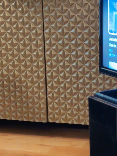 The image shows a close-up view of a section of furniture or built-in cabinetry featuring a repeating geometric pattern of raised, three-dimensional pyramids. The surface is a consistent golden-brown color. A television screen is partially visible at the top left, displaying the words "Jennifer" and "Live in," with a blue background. A dark border of the television is also visible. To the right of the patterned surface, a section of light-colored wood, possibly part of a frame or wall, is also visible. There is no context provided in the image to determine the exact location in Seattle.