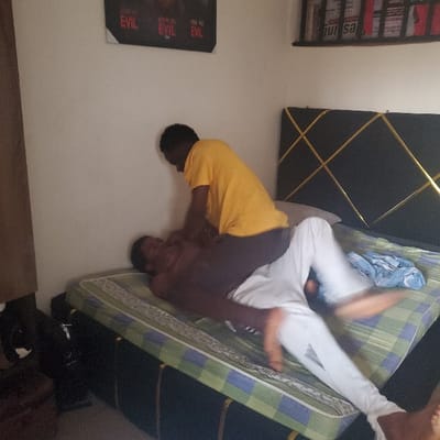 This image captures an interior scene in Eziobodo, Nigeria. Two individuals are on a bed with a black headboard featuring a gold geometric line pattern and a mattress covered by a light green and blue plaid sheet.
One individual, wearing a yellow t-shirt and dark trousers, is positioned kneeling or crouching over another individual. The second individual is shirtless, wearing white trousers, and is lying supine on the mattress. The person in the yellow t-shirt is leaning forward, with hands placed on or near the chest and upper abdomen area of the individual beneath. The lower individual's legs are elevated and display motion blur, indicating recent or ongoing movement. A crumpled light blue garment rests on the mattress near the lower individual's legs.
The background consists of a white wall. A black poster with red text, including phrases like "SEE NO EVIL," is affixed to the wall. Above the headboard, a shelf holds several books or magazines, one clearly displaying "BURSA" on its spine. To the left of the bed, a dark, indistinct object, possibly furniture, is visible.