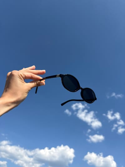 A low-angle, medium shot depicts a tan-skinned human hand holding a pair of black, round sunglasses against a clear, bright blue sky dotted with white cumulus clouds. The hand appears to be holding the sunglasses by one of the temples. The sunglasses are the focal point, positioned in the center of the frame. The location is indeterminate, but given the prompt, it is presumed to be in Valle de Bravo, Mexico. No other people or objects are visible in the frame.
