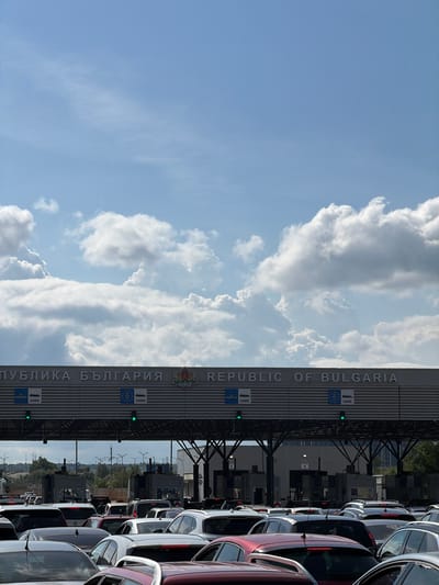 The image depicts a border crossing into Bulgaria. A large sign reading "ПУБЛИКА БЪЛГАРИЯ REPUBLIC OF BULGARIA" is visible. The sign has the EU flag and the Bulgarian coat of arms on it. There are many cars lined up in what appears to be a queue. The image was taken at the Kapitan Andreevo border crossing point. The sky is visible with many white clouds. Green traffic lights are lit.