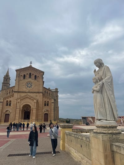 The image depicts the Basilica of the National Shrine of the Blessed Virgin of Ta' Pinu in Għasri, Gozo, Malta, under an overcast sky. The large, light-brown stone basilica occupies the background, featuring a central arched entrance, a rose window, a prominent dome, and a bell tower on its left side. A cross surmounts the main facade.

In the right foreground, a tall white stone statue stands on a classical pedestal, positioned atop a low stone wall. The statue portrays a robed adult figure with one hand raised and the other resting on the shoulder of a smaller, robed child figure standing beside them.

A paved plaza extends from the basilica's entrance towards the viewer, displaying a pattern of grey and reddish-brown sections. Numerous individuals are present in the plaza. In the immediate foreground, two young adults walk towards the camera: a female on the left in a dark jacket and blue jeans, and a male on the right in a light blue cardigan, dark pants, and a grey shoulder bag, looking directly at the camera. Multiple other people are scattered across the mid-ground, walking towards or away from the basilica entrance. To the far right, beyond the base of the statue, two individuals are seated on a bench, with a section of decorative mosaic art visible on a wall further in the background. The sky is uniformly grey and heavily clouded.