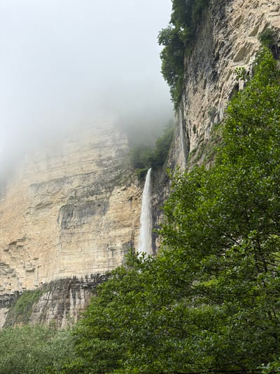 A tall, thin waterfall cascades down a sheer, beige rock face on a foggy day in Kinchkhaperdi, Georgia. The cliff is layered with horizontal striations and patches of dark, wet rock, with dense green foliage growing from its upper edge and in a thick cluster in the foreground on the right. The sky is obscured by thick, white fog, which partially envelops the upper portion of the waterfall and cliff. The lower part of the image shows more green trees and bushes.
