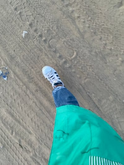 A person stands on a sandy surface, wearing blue jeans and a light grey canvas shoe. Their right foot is visible. To the right, a portion of a green garment, likely a shirt or jacket, is visible with a white striped pattern. The sand appears coarse and uneven with tire tracks visible. The lighting is bright.