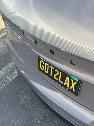 The image shows the rear of a grey Tesla vehicle parked on asphalt. The Tesla logo is visible on the trunk, along with a carbon fiber spoiler. A yellow license plate with black text reads "GOT2LAX." The license plate also has a smaller sticker with the text "J 1752566." The scene appears to be outdoors, in what looks like a parking lot. The lighting suggests it is daytime. There are no people visible in the image.