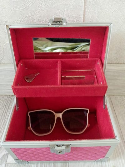 An opened, pink and silver, quilted jewelry box sits on a light wood floor against a textured white wall. The interior of the box is lined with red velvet. The top compartment has a mirror, a small silver key, and several narrow compartments. The lower compartment contains a pair of light-colored sunglasses. The scene is static. There is no indication of the location being Prigorognoye, Kyrgyzstan.