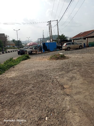The image shows a view of a street in Ado Ekiti, Nigeria, at the coordinates 7.639 latitude and 5.202 longitude. The street is paved with gravel and there are several power lines overhead. There are two cars parked on the right side of the street. There is a small blue building behind the cars on the right, and a large blue building behind the cars on the left. There are several people in the distance, but they are too far away to be identified. There are also several trees in the background. The view from the camera is from the edge of the street, looking toward the center of the street and the buildings beyond. The scene is generally quiet and peaceful. The weather is sunny and clear.