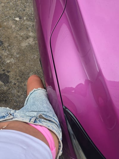 A high-angle, close-up shot shows the lower body of a person wearing a white t-shirt, distressed denim shorts, and pink underwear. The person is standing next to a shiny, metallic pink car. The background is a rough, gray concrete surface. The image is shot outdoors, and the lighting suggests daytime. The location is identified as Ban Yid, Thailand.