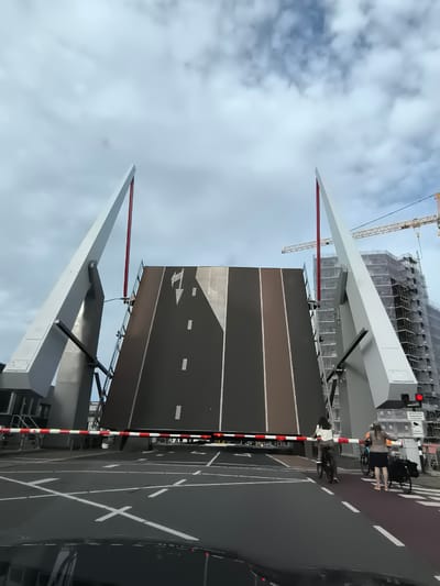 The image shows a bascule bridge in Alkmaar, Netherlands, raised to allow a boat to pass underneath. The bridge deck is dark grey with white lane markings, and it is supported by two large, white, angular structures. A red and white striped barrier arm is lowered across the road, indicating that the bridge is open. Two people are visible on bicycles on the right side of the road, waiting for the bridge to lower. In the background, a building under construction is visible, with scaffolding and a yellow crane. The sky is overcast with some clouds.