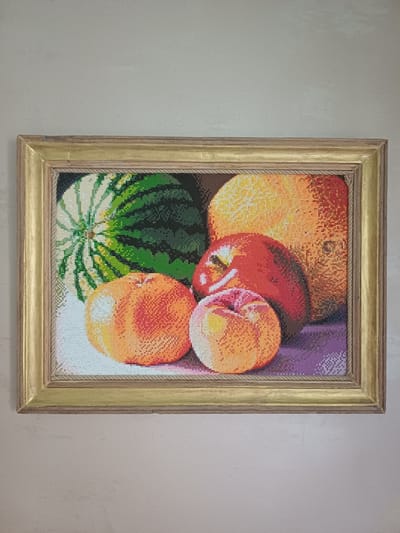 A framed still life artwork depicts a variety of fruits, including a watermelon, an orange, and two apples, arranged on a purple surface. The artwork is a mosaic of small, colored squares, giving it a pixelated appearance. The frame is made of wood and has a gold finish with a rope-like texture along its inner edge. The artwork is hanging on a textured, light-colored wall. The scene appears to be indoors, possibly in a home or gallery, located in the city of Chaika, Ukraine.