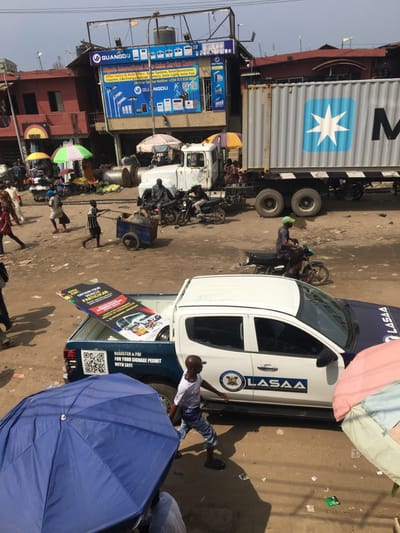 An outdoor scene shows a busy street with a mix of vehicles, pedestrians, and market activity. A large shipping container truck dominates the left side of the frame, parked near a building displaying "GUANGDU" signage. To the right, a white LASAA vehicle is parked with its rear door open, displaying a sign about signage permits. People are gathered around the vehicle. In the foreground, part of a blue umbrella is visible, suggesting a vendor's stall. The overall impression is a bustling commercial environment in a developing area.