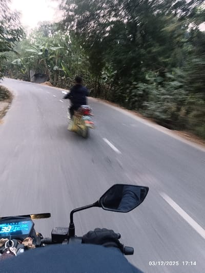 The image depicts a first-person view from a moving motorcycle in Kaliganj, Bangladesh, at 17:14 on March 12, 2025. In the foreground, the handlebars, digital dashboard displaying "38," and a rearview mirror of the photographer's motorcycle are visible. A gloved hand holds the right grip, and a dark blue garment covers the rider's lap. Keys with a small brown charm hang from the ignition area. The right mirror reflects a portion of the rider's leg, clad in blue.
The midground features an asphalt road with a white dashed line, curving slightly to the left in the distance. Ahead, another individual rides a blurred, yellow-green scooter, also in motion, wearing a dark jacket. The background consists of dense, green foliage, including trees and prominent banana plants, lining both sides of the road, suggesting a rural or semi-rural environment. The lighting indicates late afternoon conditions.