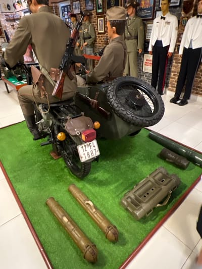 A diorama showcases a World War II-era motorcycle with sidecar and weaponry, possibly a German model. Two soldier mannequins are placed near the motorcycle, one carrying a rifle. Several military objects are present, including a binocular case, two rocket launchers, and a metal tube. The diorama is set on green artificial turf and appears to be part of a larger historical display of military uniforms and artifacts. The license plate on the motorcycle reads "12-F4 1887".