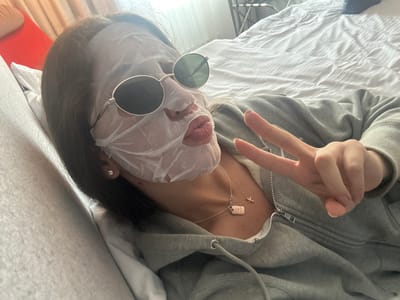 An individual is captured in a close-up, high-angle shot, lying down on a bed. The person is wearing a white sheet face mask covering most of their face, along with dark, round-framed sunglasses that rest over the mask. Their lips are pursed in a "duck face" expression, visible through the mask's mouth opening. A dark-haired individual, they are clad in a grey zippered hoodie. With their right hand, they are giving a peace sign (V-sign).

The setting appears to be a bedroom, with a rumpled white bedspread or duvet covering the bed. Bright natural light streams in from a window in the background, partially obscured by light-colored curtains or blinds, indicating it is daytime. A red surface, possibly a headboard or pillow, is visible on the left side of the frame.

Notable accessories include a small, sparkling stud earring on the person's visible left earlobe, and two silver necklaces. One necklace features a small, rectangular pendant, while the other holds a tiny cross charm. The overall tone of the image suggests a moment of casual relaxation and self-care, possibly accompanied by a playful mood. No discernible text is visible on any objects in the image. The context given places this scene in Dortmund, Germany.