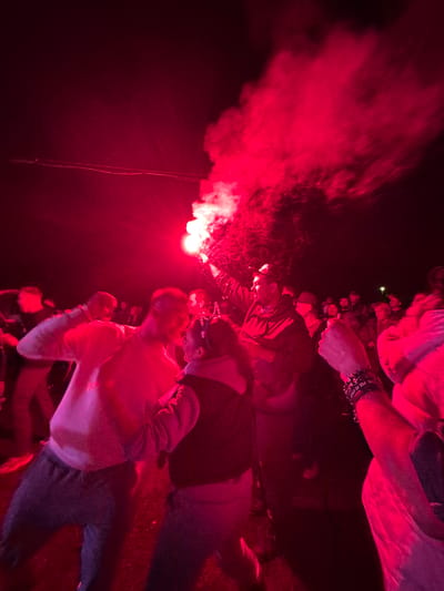 The image depicts an outdoor nighttime gathering of multiple individuals in Большая Кивара, Russia, illuminated predominantly by the intense red light and smoke emitted from a handheld flare. A male individual positioned towards the right of the frame holds the actively burning red flare aloft in his right hand, generating a thick plume of red smoke that dissipates into the dark sky. This illumination casts a strong red hue over the entire scene and the attending crowd.
In the mid-ground, to the left of the flare-holder, another male individual wearing a light-colored sweatshirt is visible, with his left arm raised or bent, his body oriented towards the activity. Further left and partially obscured, additional figures are present. In the lower-left foreground, a person in a light top and dark pants is in motion. In the lower-right foreground, a raised arm adorned with multiple dark, possibly beaded bracelets, is visible. The background reveals a dense crowd of people, mostly silhouetted or dimly lit by the red light, indicating a significant number of participants. Overhead in the upper-left, faint horizontal lines, possibly wires or cables, are discernible against the dark sky. The ground beneath the individuals appears to be an uneven outdoor surface.