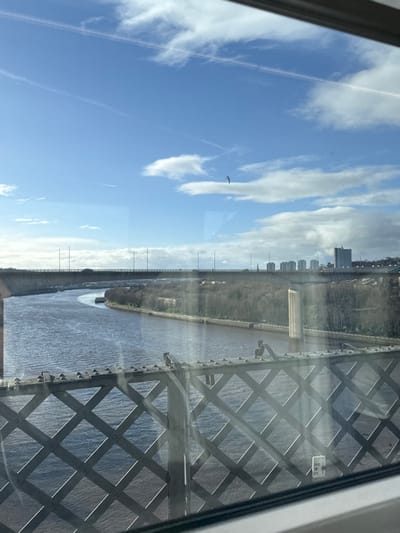 A high-angle view from a window showcases a landscape featuring a river winding between grassy banks, a city skyline in the distance, and a bright blue sky with scattered clouds. The architectural framework of a bridge dominates the right side of the image, while a bird soars freely in the sky. The scene is bathed in natural light, creating a tranquil and expansive vista.