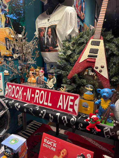 The image features an eclectic collection of items.  A red glitter-covered, V-shaped guitar ornament is intertwined with green foliage.  A t-shirt with a print featuring David Bowie and Bing Crosby lays underneath. A red "ROCK-N-ROLL AVE" sign rests among various figurines and toys, including a blue bear and a red character with a wreath.  A "Build Your Own Rock Guitar" kit is also visible, against a musical-themed background with piano keys and music notes.