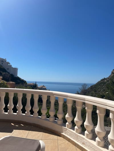 The image shows a view from a balcony overlooking the sea. The location is likely in or near El Poble Nou de Benitatxell/Benitachell, Spain. In the foreground is a white balustrade of a balcony. Behind the balcony are the clear blue sea and sky. On either side of the sea are rocky coastlines covered with vegetation. A glimpse of building roofs is visible along the coastline. A sun lounger is partially visible on the patio.
