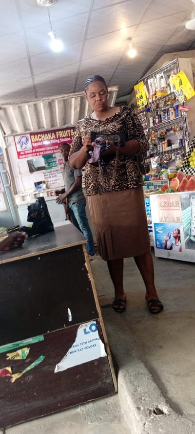 This image depicts a scene inside a shop, likely a convenience store or market stall, in Aviation Quarters, Nigeria. The setting appears to be indoors with a tiled ceiling and fluorescent lighting, suggesting it's daytime.

The primary subject is a woman with braided hair, wearing a patterned long-sleeved shirt and a brown skirt, standing and holding a small bag and a mobile phone. She is looking down at her hands. To her left, a younger man in a grey t-shirt and jeans is visible, looking towards the camera with a neutral expression. In the foreground, a dark wooden counter is visible, with a black plastic bag on it. Behind the counter, shelves are stocked with various goods, including toiletries, keys, and some fruit. A sign for "BACHAKA FRUITS" is prominent on the wall, indicating the type of business. Other visible text on signs includes "AKBAR," "LO," and "NEW CAP COLORS," suggesting a variety of products are for sale. The overall atmosphere seems to be one of everyday commerce.
