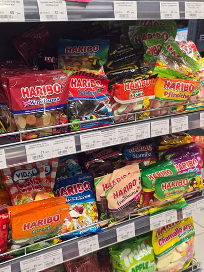 The image shows a retail shelf in a store, likely a supermarket or convenience store, stocked with various bags of Haribo gummy candies. The candies are arranged in rows on metal shelves, with price tags visible for some items. The lighting is bright and consistent with indoor store lighting. The products are predominantly bags of Haribo gummies, with labels in Ukrainian and German. Some of the visible product names include "Haribo Wine Gums," "Haribo Salino," "Haribo Wasser Melonen," "Haribo Pfirsiche," "Haribo Color-rado," "Haribo Starmix," "Haribo Quaxi," "Haribo Fruitmania," and "Haribo Chamallows Rombls." The price tags indicate prices in Ukrainian Hryvnia (грн.). The environment suggests a commercial setting focused on confectionery sales. There are no people visible in the image. The location mentioned, Petropavlivska Borshchahivka, Ukraine, is a town, suggesting this is a retail outlet within that area.