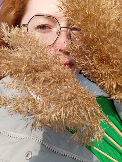 A close-up, slightly canted photograph shows a person with red hair and glasses partially obscured by dried reed stalks. The person's left eye is visible, along with part of their cheek and lips. They wear black-rimmed glasses. The reed stalks are light brown and feathery. Behind the reeds and person, there's a glimpse of a light green object with linear features, and part of a light gray or white garment with a zipper and button detail. The photograph was taken in Yurgamysh, Russia.