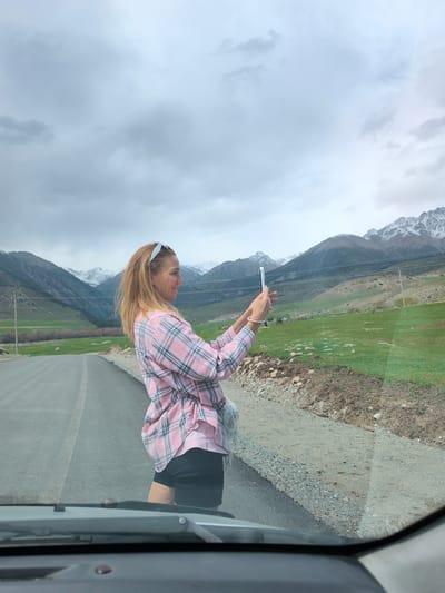 The image shows a woman taking a picture with her smartphone on a road in Semenovka, Kyrgyzstan. She is wearing a pink and black plaid shirt, black shorts, and a white headband. She is standing on the side of the road, with a landscape of green fields and snow-capped mountains in the background. The sky is overcast with gray clouds. The image is taken from inside a vehicle, with the dashboard and windshield visible at the bottom of the frame.