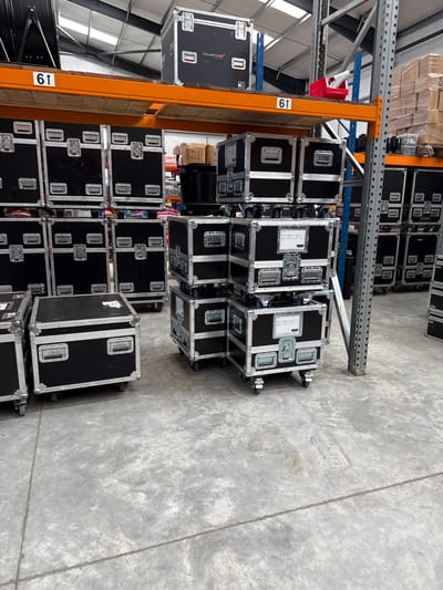 The image depicts the interior of a warehouse, likely in an industrial area. The location is specified as Epping Forest, United Kingdom. The warehouse is organized with metal shelving units, identified by the number "61" on an orange support beam. Stacks of black, rectangular cases with metal trim and wheels dominate the scene. These cases appear to be for transporting equipment, potentially audio-visual or musical instruments, given the presence of handles and latches. The cases are neatly arranged on the floor, some stacked on top of each other. The warehouse floor is a light gray concrete. There are no visible people in the image. The lighting is bright, suggesting the presence of large windows or artificial lighting fixtures. Pallets of boxes are stacked on the shelving units, further indicating the purpose of the building.