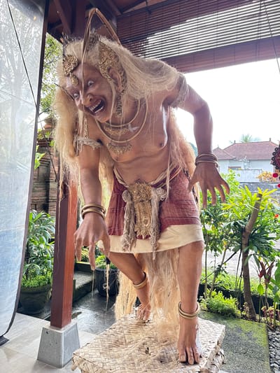 A full-body, eye-level, medium shot captures a life-sized, theatrical effigy of a mythical Balinese creature, likely a Rangda or Barong, poised in an outdoor setting. The creature, with its wide-open mouth and disheveled white hair, appears to be mid-action, leaning forward with its hands outstretched. It wears traditional Balinese attire, including a red and gold skirt, ornate gold jewelry around its neck, arms, and head, and a gold sash. The effigy is standing on a woven mat, which is elevated on a wooden and concrete pedestal. In the background, lush green foliage and Balinese architecture, including a brick wall and tiled roofs, are visible, suggesting the location is a garden or courtyard in Payangan, Indonesia. The lighting suggests it's daytime, with bright sunlight illuminating the scene.