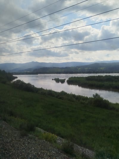 The image depicts a serene natural landscape under a cloudy sky, likely taken in Solnechnyy, Russia. The scene is dominated by a wide, calm river flowing through a valley. Rolling, tree-covered hills form the background, fading into a misty, mountainous horizon. Several small, vegetated islands dot the river's surface, breaking the expanse of water.

In the foreground, a grassy slope leads down to the riverbank, with some gravel and sparse yellow wildflowers visible at the very bottom left. The overall impression is one of tranquility and natural beauty. The time of day appears to be late afternoon or early evening, as suggested by the diffused light and overcast conditions. The weather is cloudy, but not stormy, contributing to the subdued atmosphere.

Several power lines stretch across the upper part of the frame, creating a geometric contrast with the organic shapes of the landscape. There is no visible text in the image. The scene evokes a sense of peacefulness and the vastness of nature.