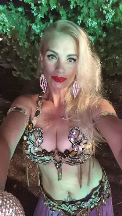 This is a selfie of a blonde woman wearing a jeweled belly dancing costume. The costume includes a bra adorned with rhinestones and beads, a jeweled belt, and what appears to be a sheer purple skirt. She is also wearing large, diamond-shaped earrings and armbands. Her makeup is dramatic, with bold red lipstick and winged eyeliner. Her expression is confident, with a direct gaze at the camera. The background is a blur of dark green foliage, suggesting an outdoor setting, possibly at night or in low light, as there are hints of artificial illumination on the leaves. The woman's blonde hair is long and styled in loose waves. The overall impression is that she is dressed for a performance or a special occasion. While the exact location is not explicitly visible, the provided context indicates Kuşadası, Turkey, which is a popular tourist destination known for its cultural performances and vibrant nightlife.