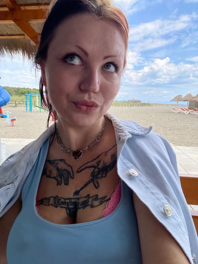 A woman with tattoos is posing for a selfie. She is wearing a blue tank top and a light blue denim jacket. She also has a silver necklace with a red flower pendant. The background appears to be a beach scene, with sand, the Adriatic Sea, and sunbeds. It looks like Velika Plaza Beach in Ulcinj, Montenegro.