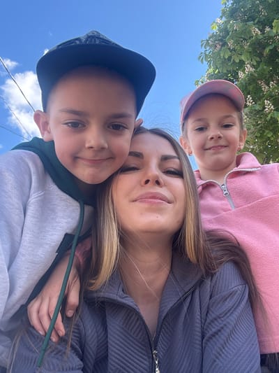 A selfie features a woman with long brown hair and two children. The woman is in the middle, wearing a dark gray ribbed jacket. A young girl with a pink cap and a pink jacket is positioned above her, and a young boy with a dark cap and a white sweatshirt is located below her. The background reveals a blue sky with some trees and what appear to be power lines. No information suggests this photo was taken in Slobodka, Russia.