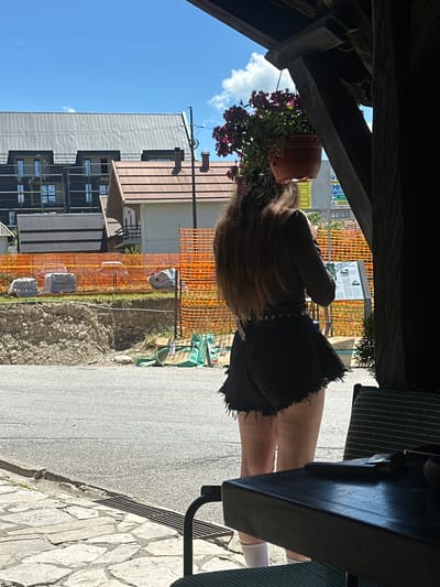 A woman with long brown hair stands in the foreground, facing away from the camera. She is wearing a dark, off-the-shoulder top and distressed black denim shorts. A silver-buckled belt is visible around her waist. She is standing on a stone-paved area next to a road. In the background, there is a construction site with orange safety netting, stacks of building materials, and several buildings under construction or recently built. The sky is blue with some scattered clouds. The location is identified as Dulovine, Montenegro.