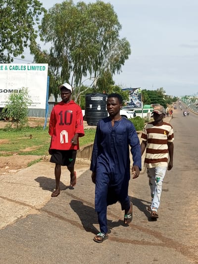 Three young men are walking on a paved road in Garko, Nigeria. The man on the left is wearing a red hoodie with the number "10" prominently displayed on the front, black shorts, and red flip-flops. He is also wearing a white baseball cap. The man in the middle is wearing a dark blue, long-sleeved tunic and dark pants, and he is barefoot. The man on the right is wearing a brown and white horizontally striped t-shirt, light-colored ripped jeans, and a baseball cap. He is also barefoot. In the background, there are trees, a large black water tank, a billboard with text and graphics, and a busy street with traffic, including cars and motorcycles. The sky is partly cloudy.