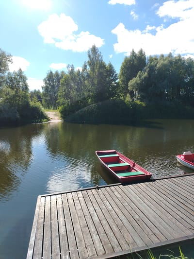 The image depicts a calm, sun-drenched scene at a waterfront location, likely a park or recreational area. In the foreground, a weathered wooden pier extends into a dark, reflective body of water. Two small, red rowboats with green seats are docked at the pier. The boat closer to the foreground is fully visible, while the other, partially obscured, has the brand name "SAVA" visible on its stern. The water's surface is gently rippled, reflecting the blue sky and scattered white clouds. The background features a lush, green treeline with a sandy incline leading down to the water on the left side of the image. The sky is a vibrant blue, with bright sunlight casting highlights and shadows. The scene is devoid of human presence. This image was captured on August 10, 2023, at the "Belaya Gora" park in the city of Stroitel', Belgorod Oblast, Russia.