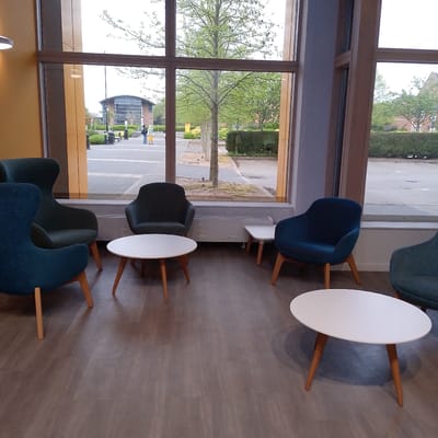 This image captures a modern interior space, likely a lounge or waiting area, with large floor-to-ceiling windows offering a view of an outdoor pedestrian zone. The indoor area features light wooden-look laminate flooring and warm yellow and grey walls. Furnishings include several mid-century modern style armchairs upholstered in dark teal and dark grey fabrics, all with light wooden legs. Two oval white coffee tables with matching wooden legs are placed between the chairs. A low, white radiator unit runs along the base of the windows.
Through the expansive windows, an outdoor paved area is visible, typical of a campus or public square in Coventry, United Kingdom. The weather appears overcast, providing soft, diffused lighting. Several trees with fresh, light green leaves, indicative of spring or early summer, line the paved walkway. A row of small birds can be seen on the ground near the trees. In the mid-distance, two people are present on the paved area: one dressed in dark clothing with a backpack, and another in a light yellow dress and hat. They appear to be walking or standing. Further in the background, a modern building with a curved roof and extensive fenestration is visible on the left, while on the right, residential or institutional buildings are partially obscured by hedges and trees. No discernible text is visible in the image.