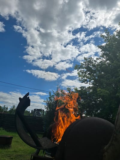 In Ермолаево, Russia, under a bright blue sky dotted with white, fluffy clouds, a fire burns vigorously in a metal structure, likely a grill or brazier. The flames, a vibrant orange and yellow, leap upwards. In the foreground, a dark, curved metal object obscures a portion of the fire and the surrounding scene. To the left of the fire, a dark silhouette of a hand with an extended index finger is visible against the sky. In the background, a dark wooden fence runs horizontally, and behind it, a green tractor is parked on grassy terrain. Trees with lush green foliage flank the scene on the right and in the distance. A dark, cylindrical object, possibly a barrel, rests on the grass to the left. Power lines are strung across the sky.