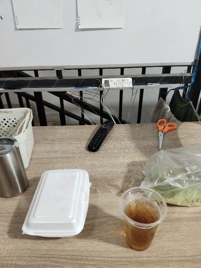 A wooden table with a white styrofoam container, a clear plastic cup with brown liquid, a metal thermos, a remote control, a pair of scissors, and a plastic bag with green leafy vegetables. The table is located in a home in Telukjambe, Indonesia, at the coordinates -6.308 latitude and 107.282 longitude. Hanging above the table is a metal railing with several wire hangers hanging on it. The scene appears to be casual, suggesting a living space in the home.