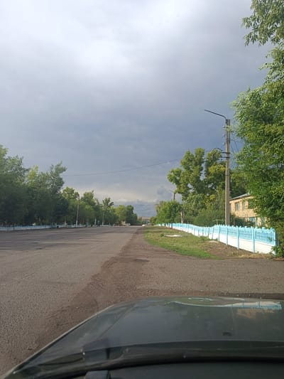 This is a photograph taken from the perspective of a vehicle looking down a paved road in an outdoor setting. The road appears to be in a town or village, with a line of trees bordering the left side and a light blue and white decorative fence lining the right. Beyond the fence, a building with a light-colored brick facade is visible. A power pole with electrical wires stands near the fence, and more trees are scattered throughout the scene. The sky is predominantly overcast with heavy, dark clouds, suggesting impending rain or recent precipitation. The overall mood is somewhat somber due to the weather, but the greenery of the trees and the clear visibility indicate it is daytime. The reflection on the car's hood suggests the photo was taken in the morning or afternoon. No people are visible in the frame. The image captures a typical street scene in a rural or semi-urban environment under a stormy sky. The location is identified as Молодежный, Kazakhstan.