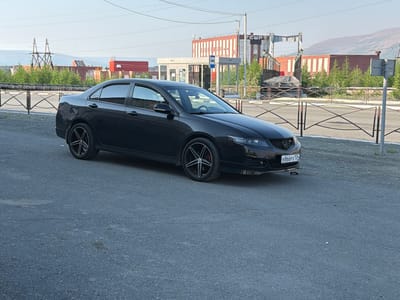 A black sedan is parked on an asphalt surface. The car is facing towards the right of the frame, with its front end slightly angled away from the viewer. It has custom black rims with a five-spoke design. A license plate with the inscription "B866PX 124 RUS" is visible on the front of the car.
In the background, there are several industrial buildings with red brick and white siding. A bus stop shelter with glass panels is situated to the left of the car. Power lines and towers are also visible in the distance, along with some sparse green trees and bushes. The sky is clear and appears to be either early morning or late afternoon, given the soft light. The overall scene suggests an outdoor urban or semi-industrial setting, possibly in a northern climate given the location context of Norilsk, Russia. No people are visible in the image. The atmosphere appears calm and still.